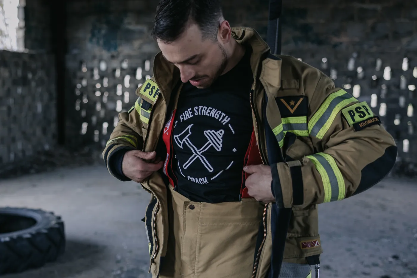 Paweł Kokot w mundurze strażaka, otwiera kurtkę, pod nią koszulka Fire Strength Coach